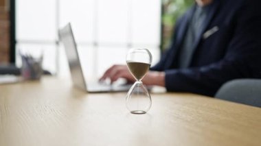 Genç İspanyol iş adamı ofisinde kum saatiyle dizüstü bilgisayar kullanıyor.