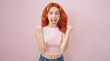 Genç, kızıl saçlı, kendine güvenen bir kadın, pembe arka planda, kazanan yüz ifadesiyle ayakta duruyor.