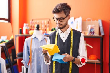 Young hispanic man tailor smiling confident holding cloths at atelier