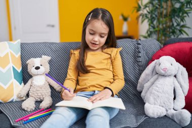 Adorable hispanic girl drawing on notebook sitting on sofa at home