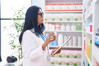 Orta yaşlı İspanyol kadın eczacı elinde ilaç şişesiyle eczanede doküman okuyor.