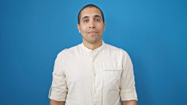 Soyutlanmış mavi arka planda ciddi bir ifadeyle bir bankta oturan İspanyol genç adam.