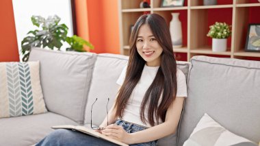 Young chinese woman reading book sitting on sofa at home