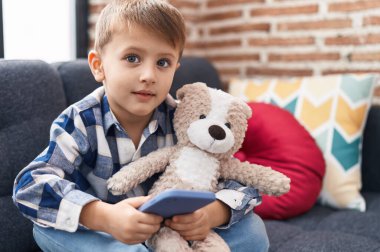 Beyaz çocuk oyuncak ayıya sarılıp evde akıllı telefon kullanıyor.