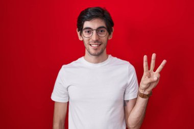 Young hispanic man standing over red background showing and pointing up with fingers number three while smiling confident and happy. 