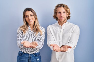 Mavi arka planda duran genç çift avuçlarını birbirine uzatmış gülümsüyor ya da jest yapıyor. Bekleme ve koruma 