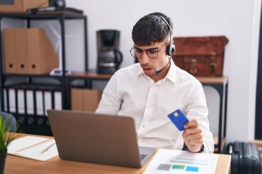 Genç İspanyol bir adam bilgisayar kullanıyor. Elinde kredi kartı var. Şok olmuş, şüpheci ve alaycı görünüyor. 