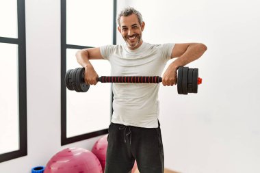 Orta yaşlı, gri saçlı, kendine güvenen, spor merkezinde ağırlık çalışması yapan bir adam.