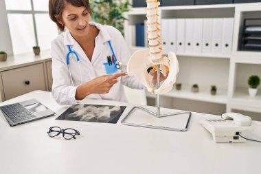 Young beautiful hispanic woman doctor pointing with finger to anatomical model of vertebral column at clinic