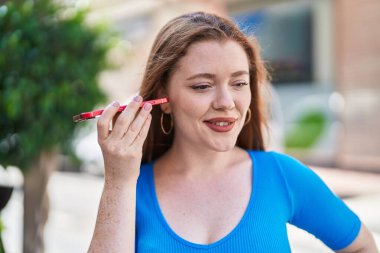 Young redhead woman miling confident listening audio message by the smartphone at street