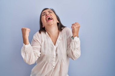 Mavi arka planda dikilen orta yaşlı İspanyol kadın kolları havada ve gözleri kapalı bir şekilde başarıyı kutluyor ve şaşırıyor. Kazanan konsept. 