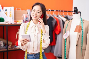 Young chinese woman tailor talking on smartphone reading notebook at atelier