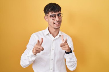 Young hispanic man standing over yellow background pointing fingers to camera with happy and funny face. good energy and vibes. 