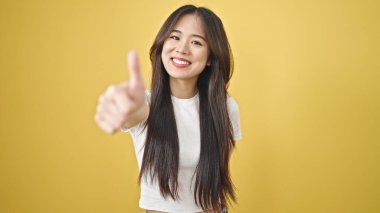 Genç Çinli kadın gülümsüyor. Kendine güveni tam. Sarı arka planda baş parmağıyla işaret veriyor.