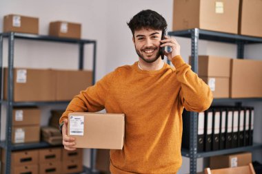Young hispanic man ecommerce business worker talking on smartphone holding package at office