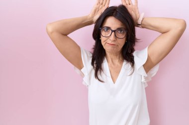 Orta yaşlı İspanyol kadın, günlük beyaz tişört ve gözlük takıyor. Tavşan kulağı hareketi yapıyor. Avuçlarında alaycı ve şüpheci bakışlar var. Paskalya tavşanı konsepti. 