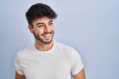 Hispanic man with beard standing over white background looking away to side with smile on face, natural expression. laughing confident. 