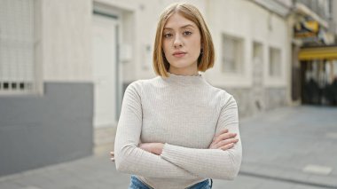 Ciddi bir ifade ve kollarıyla caddede duran genç sarışın kadın.