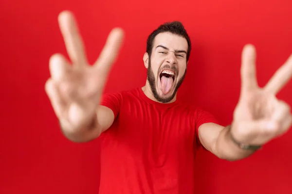 両手の指を見せて舌で微笑むカジュアルな赤いTシャツを着た若いヒスパニック系の男が勝利サインをしています — ストック写真