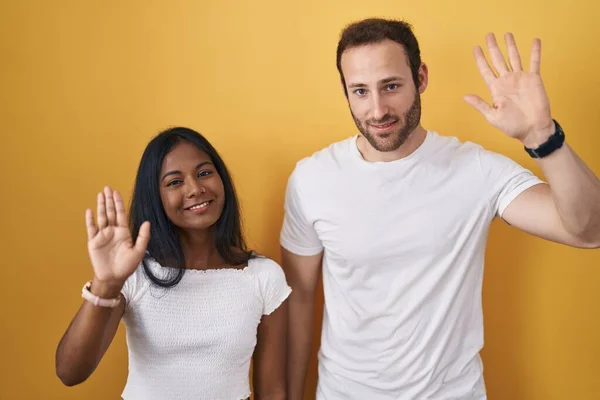 黄色の背景の上に立っている異人種間のカップルはこんにちは幸せと笑顔 優しい歓迎のジェスチャーを言う — ストック写真