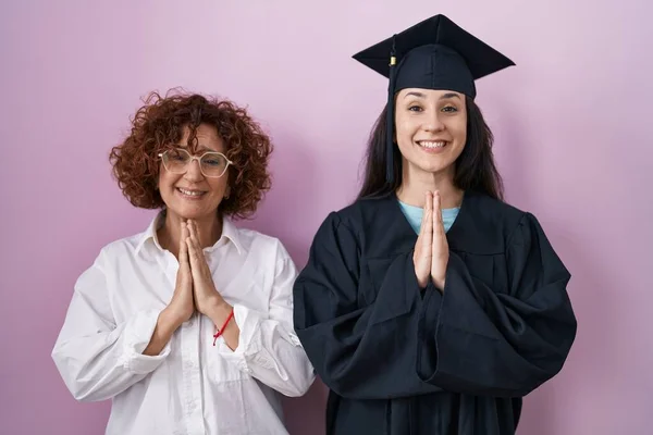 ヒスパニック系の母親と娘が卒業キャップと儀式のローブを身に着けています一緒に手で祈っています許し笑顔自信を求めて — ストック写真
