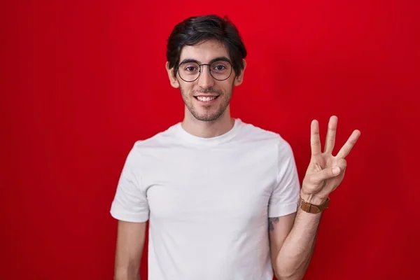 Молодой Латиноамериканец Стоит Красном Фоне Показывает Пальцем Номер Три Улыбаясь — стоковое фото