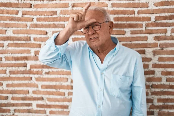 Senior man with grey hair standing over bricks wall making fun of people with fingers on forehead doing loser gesture mocking and insulting.
