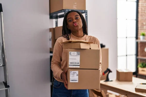 Young african american with braids working at small business ecommerce holding packages looking at the camera blowing a kiss being lovely and sexy. love expression.