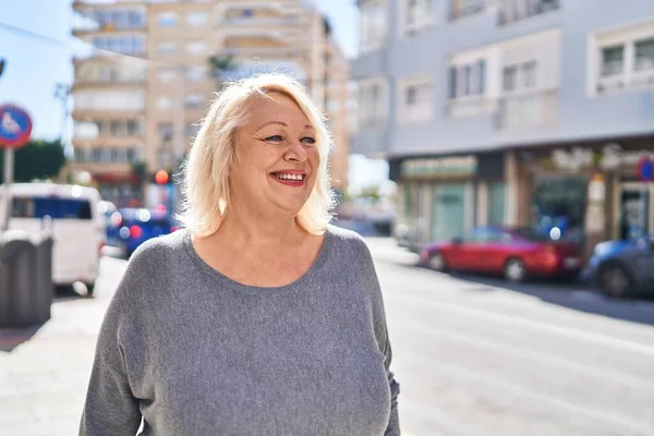 Orta yaşlı sarışın kadın sokakta kendinden emin bir şekilde gülümsüyor.