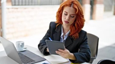 Genç kızıl saçlı iş kadını dokunmatik ped kullanıyor. Kahve dükkanının terasında not alıyor.