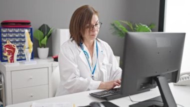 Orta yaşlı İspanyol kadın doktor klinikte bilgisayar okuyarak rapor veriyor.