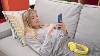 Genç sarışın kadın evdeki kanepede uzanmış müzik dinliyor.