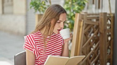 Genç sarışın kadın kahve içip kahve dükkanının terasında defter okuyor.