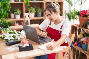 Young blonde woman florist using laptop writing on notebook at flower shop
