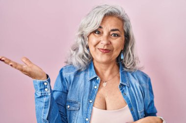 Middle age woman with grey hair standing over pink background smiling cheerful presenting and pointing with palm of hand looking at the camera. 