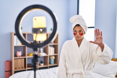 Koyu renk saçlı İspanyol kadın akıllı telefonlu güzellik dersi kaydediyor. Eli açık, dur işareti yapıyor. Ciddi ve kendinden emin bir ifadeyle, savunma hareketi yapıyor. 