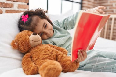 Adorable hispanic girl reading book lying on bed at bedroom