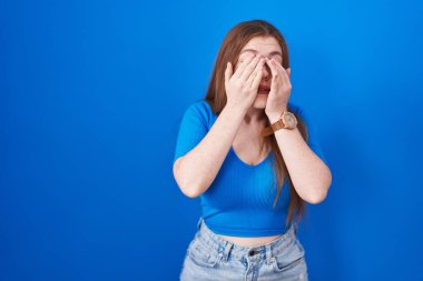 Kızıl saçlı kadın mavi arka planda duruyor. Yorgunluk ve baş ağrısı için gözlerini ovuşturuyor. Uykulu ve yorgun bir ifade. görme sorunu 