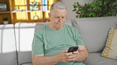 Orta yaşlı, kır saçlı, akıllı telefon kullanan adam evdeki kanepede oturuyor.