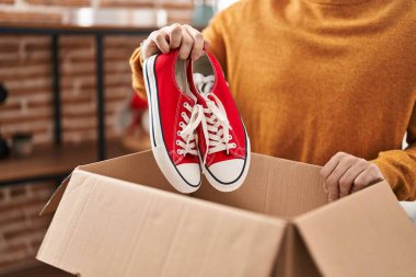 Young hispanic man smiling confident holding sneakers unpacking cardboard box at new home