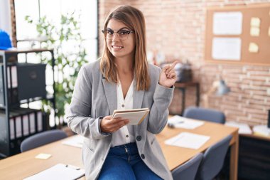 Ofiste çalışan genç İspanyol bir kadın. Gözlüklü, yüzünde kocaman bir gülümseme olan, elini kaldırıp kameraya bakan bir kadın.. 