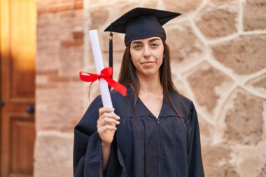 Young hispanic woman wearing graduated uniform holding diploma at university