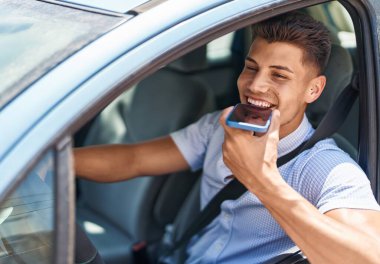 Young hispanic man talking on the smartphone sitting on car at street