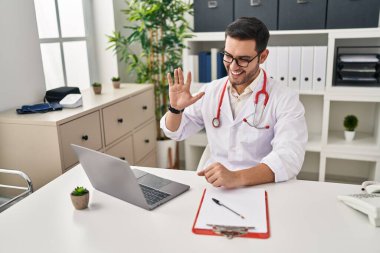 Sakallı genç İspanyol doktor klinikte online randevu veriyor. Olumlu ve mutlu görünüyor. Kendine güvenen bir gülümsemeyle dişlerini gösteriyor. 