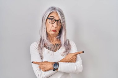 Beyaz arka planda duran gri saçlı orta yaşlı kadın her iki tarafı da parmaklarıyla işaret ediyor, farklı yönlere bakıyor. 