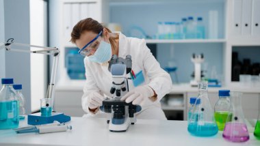 Laboratuvarda mikroskop kullanarak orta yaşlı İspanyol kadın bilim adamı üniforması ve tıbbi maske giyiyor.