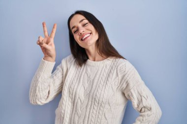 Mavi arka planda duran esmer kadın kameraya bakıp zafer işareti yapıyor. İki numara.. 