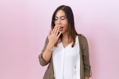 Orta yaşlı Çinli kadın pembe arka planda gözlük takıyor. Ağzı eliyle kapatmaktan sıkılmış. Huzursuz ve uykulu.. 