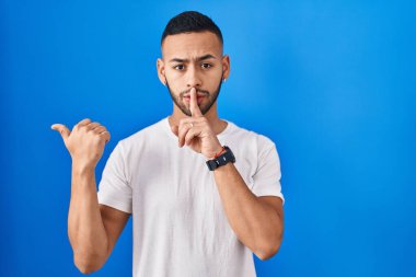 Mavi arka planda dikilen genç İspanyol adam sessiz olmak istiyor. Parmakları dudaklarında, elleri yandan işaret ederek. sessizlik ve gizli konsept. 
