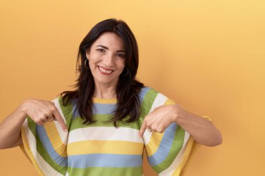 Middle age hispanic woman standing over yellow background looking confident with smile on face, pointing oneself with fingers proud and happy. 